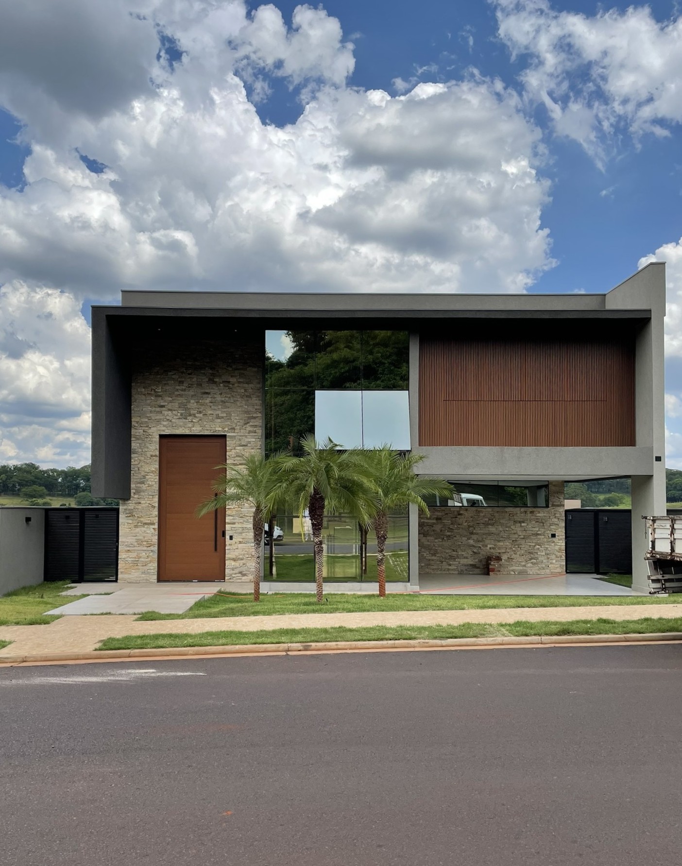Fachada contemporânea da Casa Brise com pedra, brise de madeira, porta pivotante e palmeiras sob céu azul