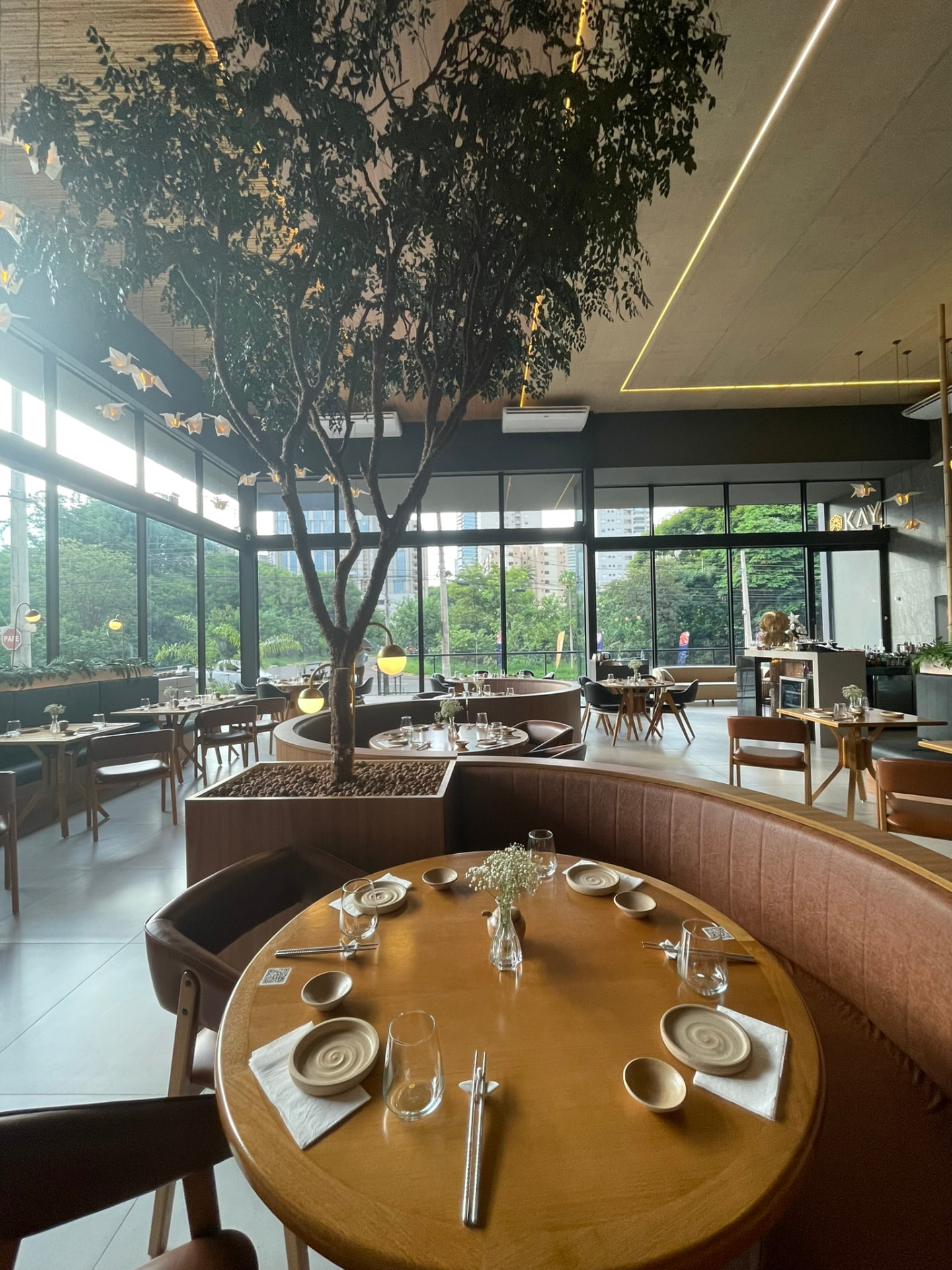 Salão do restaurante em Ribeirão Preto com árvore central, mesas redondas em madeira e tsurus iluminados suspensos