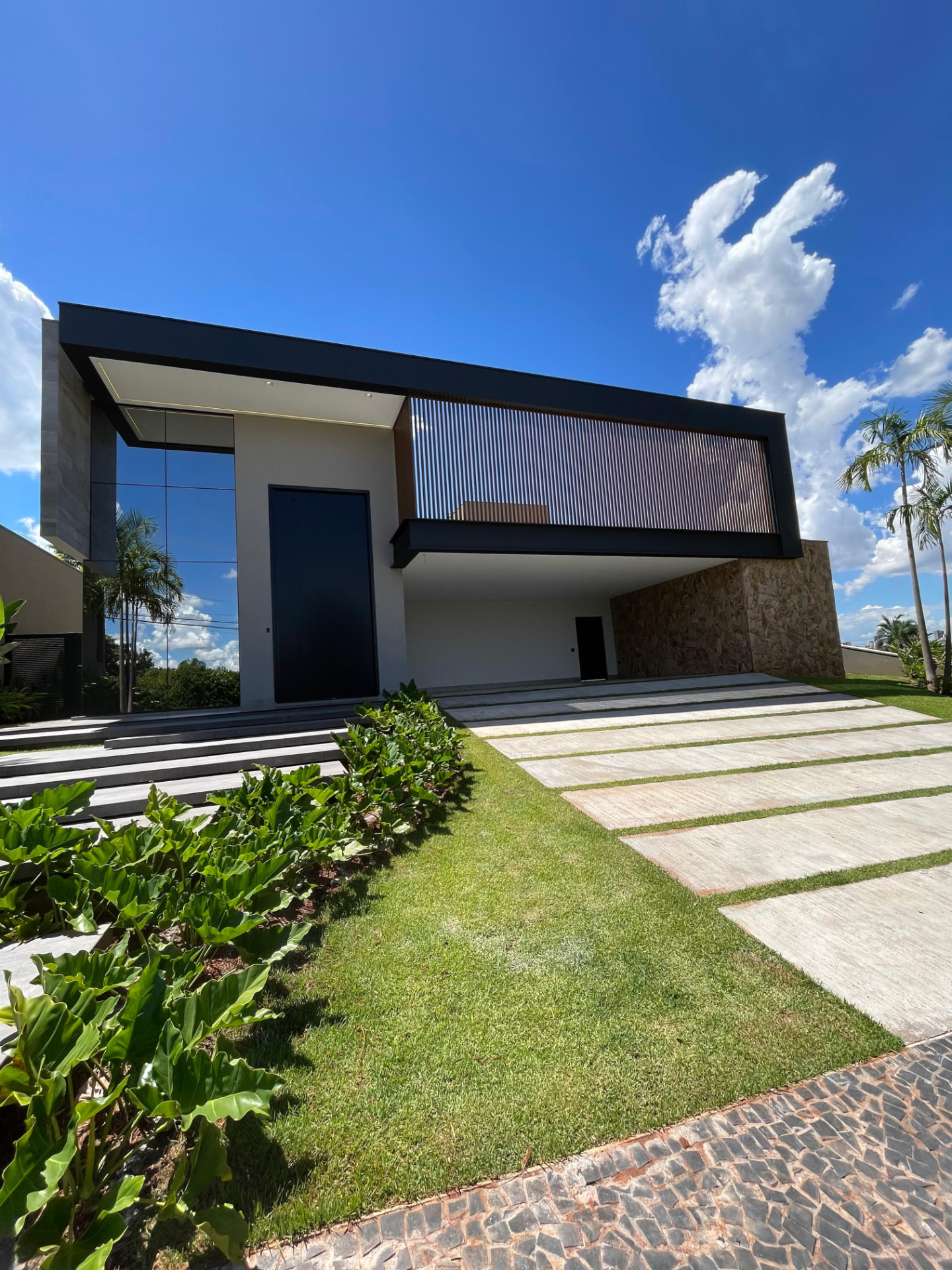 Fachada contemporânea da Residência G+G com volume suspenso preto, brise ripado de madeira e paisagismo tropical