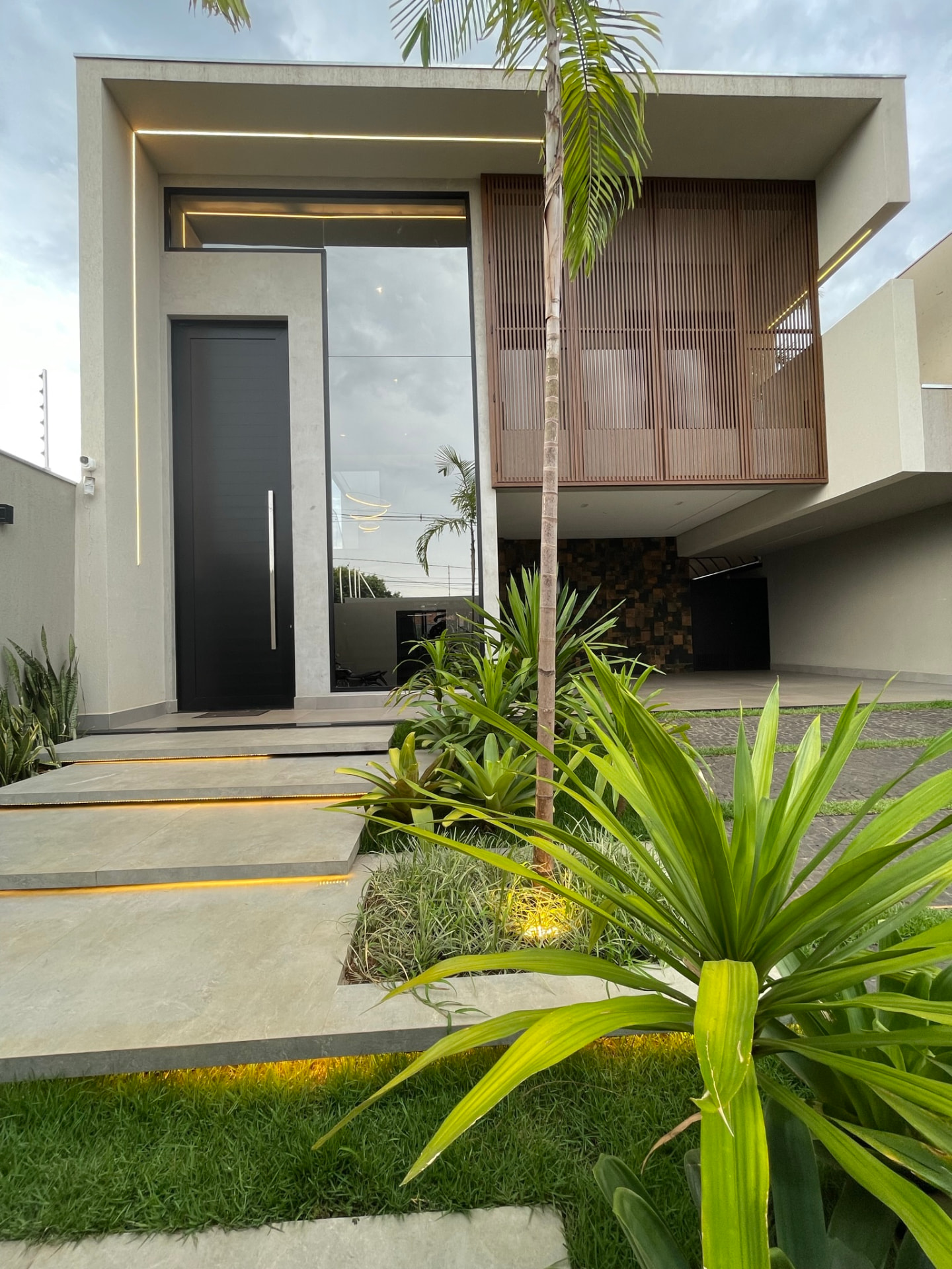 Fachada contemporânea da Residência L+M com painel ripado, iluminação linear e jardim tropical em Ribeirão Preto